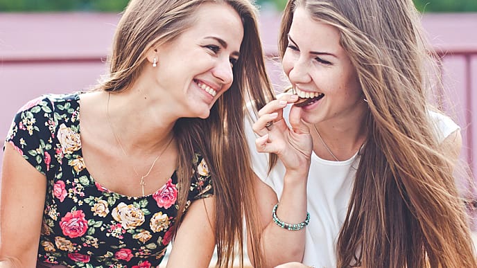 Two young female friends laugh and eat chocolate outdoors